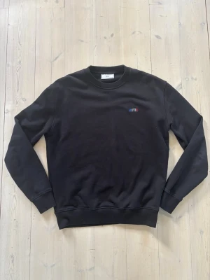 Ami Paris tröja  - Stilren svart sweatshirt från Ami Paris med liten broderad logga i blått, vitt och rött på bröstet. En liten defekt i magen på tröjan. Men inget som syns (se sista bild)😀