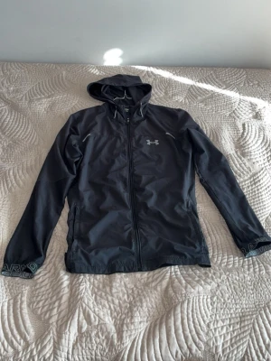 Svart vindjacka från Under Armour - Från JD sports äkta 1080 kostade den.Snygg svart vindjacka från Under Armour med huva och hel dragkedja framtill. Jackan har reflekterande detaljer och logga på bröstet samt tryck på ärmsluten. Perfekt för blåsiga dagar och sportiga outfits.