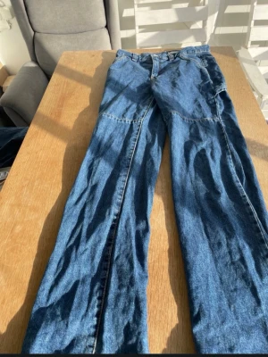 Blå raka jeans med sömdetaljer - Säljer ett par blå jeans med raka ben och markerade sömmar längs framsidan. Jeansen har klassisk femficksdesign och är gjorda i denim. Perfekta för dig som gillar en avslappnad och trendig look.