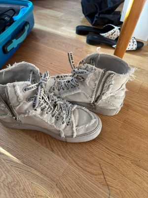 Vita high top sneakers med råa kanter - Säljer ett par beige/vita high top sneakers från Zadig & Voltaire med coola råa kanter och dragkedja på sidan. Skorna har vita snören med svart text och är gjorda i canvasmaterial. Perfekta för dig som gillar en edgy och avslappnad stil.