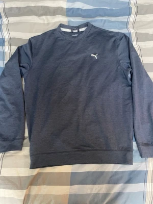 Mörkblå sweatshirt från Puma - Mörkblå sweatshirt från Puma med klassisk rund halsringning och diskret Puma-logga i vitt på bröstet. Tröjan har långa ärmar och ribbade muddar vid ärmslut och nederkant. Perfekt för en avslappnad och sportig stil.