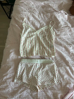 Vit pyjamas med röda hjärtan från asos - Sött pyjamasset i vitt med små röda hjärtan. Linnet har tunna axelband och spetsdetaljer vid urringningen. Shortsen har knappar framtill och elastisk midja. Perfekt för mysiga kvällar och sovmorgnar.