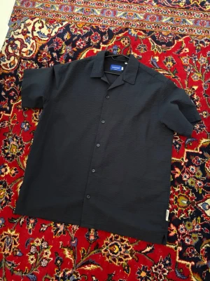 Svart kortärmad skjorta Jack & Jones - Säljer en svart kortärmad skjorta från Jack & Jones Originals. Skjortan har klassisk krage, knappar framtill och är tillverkad i ett strukturerat bomullstyg som ger en schysst känsla. Perfekt till sommaren och enkel att matcha med olika outfits.