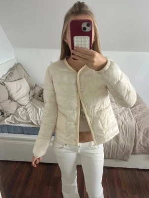 Jacka  - Snygg krämvit dunjacka med rund halsringning och tryckknappar framtill. Västen har två praktiska fickor framtill. Perfekt att slänga över en tröja för extra värme. Materialet är lätt och mjukt. Slutsåld på hemsidan i alla storlekar och är helt oanvänd med prislapp kvar! (Öppen för prisförslag)