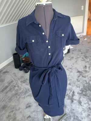 Marinblå skjortklänning från Ralph Lauren - Marinblå skjortklänning från Ralph Lauren med knappar framtill, två bröstfickor och knytskärp i midjan. Klänningen har korta ärmar och klassisk krage. Perfekt för dig som gillar en stilren och tidlös look.