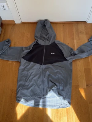 Grå och svart Nike vindjacka - Snygg vindjacka från Nike i grått med svart panel över bröstet och vit logga. Jackan har huva, dragkedja framtill och två sidofickor. Perfekt för blåsiga dagar och sportiga outfits.