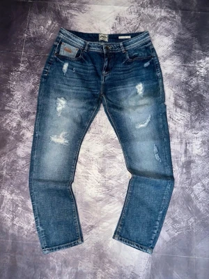 Blå slitna jeans med raka ben - Säljer ett par blå jeans med slitningar och raka ben. Jeansen har en klassisk femficksmodell och tydliga slitdetaljer framtill för en trendig look. Perfekta för dig som gillar en avslappnad och cool stil. Storlek finns på bilderna 