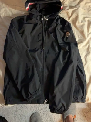 Moncler windbreaker - Snygg svart vindjacka från Moncler med dragkedja framtill och huva med rödvit rand. Jackan har elastiska muddar vid ärmslut och nederkant samt Moncler-logga på bröstet. Perfekt för blåsiga dagar och enkel att matcha med olika outfits. Använd bara 1 gång