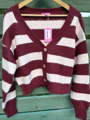 Randig burgundy kofta Edikted - Säljer en croppad kofta från Edikted i burgundy och ljusrosa breda ränder. Koftan har v-ringning, tre knappar framtill och ribbade muddar. Materialet är mjukt och fluffigt, perfekt för lager på lager och en trendig look.