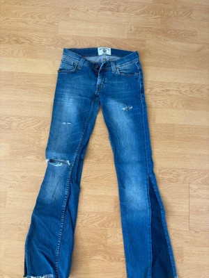 Lågmidjade Blå bootcut jeans med slitningar  - Säljer ett par blå bootcut jeans från Jeane Blush med slitna detaljer på låret och knät. Jeansen har klassisk femficksmodell, låg midja och råa påsydda kanter vid benslut. En skärphållare är trasig på en sida, men går lätt att fixa! 