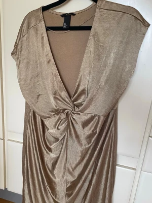 H&M omlottklänning i champagnefärg stl L - Säljer en snygg champagnefärgad klänning från H&M med v-ringning och twistad detalj framtill. Klänningen har korta ärmar och är tillverkad i ett glansigt material som ger en lyxig känsla.
