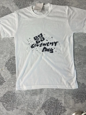 Vit Givenchy Paris t-shirt - Snygg vit t-shirt från Givenchy Paris med svart grafiskt tryck framtill.Storlek M,men känns som S.Klassisk rund hals och korta ärmar. Perfekt för dig som vill ha en clean men ändå statement look. Tillverkad i mjuk bomull för skön känsla hela dagen.