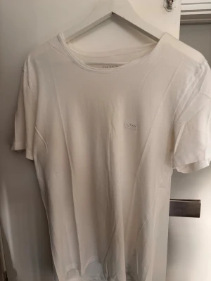 Vit t-shirt från BOSS - Säljer en stilren vit t-shirt från BOSS med diskret logga på bröstet. T-shirten har rund halsringning och korta ärmar. Tillverkad i mjuk bomull som känns skön mot huden. Perfekt basplagg för alla tillfällen.