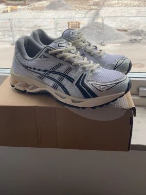 Vita ASICS sneakers med mesh och detaljer - Snygga vita ASICS sneakers med grå och svarta detaljer. Ovandelen är i mesh och syntetmaterial för bra andningsförmåga och komfort. Klassisk sportig look med ASICS-logga på sidan och vadderad häl. Perfekta för en streetwear-stil.