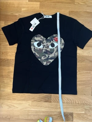 Svart Comme des Garçons PLAY t-shirt - Svart t-shirt från Comme des Garçons PLAY med stort hjärta i camouflage-mönster och ikoniskt rött hjärta med ögon på bröstet. Klassisk rund hals och korta ärmar. Perfekt statement-plagg för dig som gillar streetwear och coola prints.