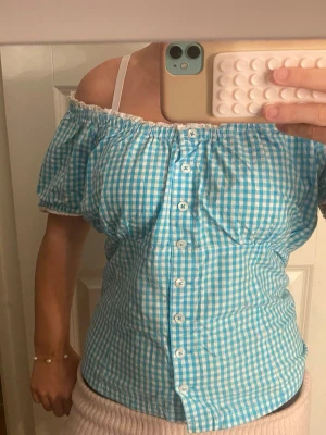 Blåvit rutig offshoulder blus y2k - Söt offshoulder blus i blåvitt rutigt mönster med korta puffärmar och knappar framtill. Blusen har en elastisk kant upptill som ger en snygg passform och är perfekt för sommarens alla tillfällen.