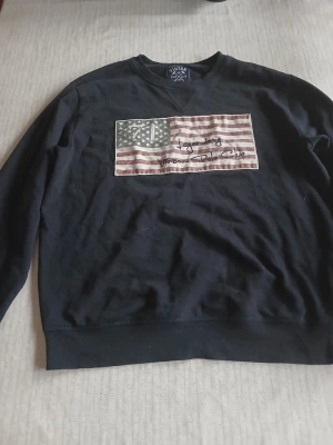 Svart sweatshirt med USA-flagga - Cool svart sweatshirt med tryck av en amerikansk flagga på bröstet och textdetaljer. Tröjan har rund hals och långa ärmar. Perfekt för dig som gillar streetwear och vill ha en avslappnad stil.