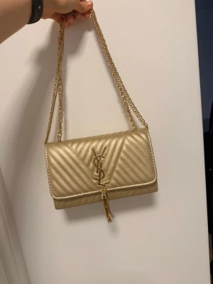 Guldig quiltad handväska med kedja - Snygg guldig handväska från med quiltat mönster och dubbla guldfärgade kedjor. Väskan har YSL-logga framtill och en dekorativ tofs. Perfekt accessoar för att lyfta din outfit. Helt ny aldrig använd.