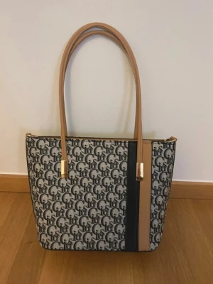 Beige och mörkblå mönstrad handväska - Snygg handväska med beige och mörkblå mönster, långa ljusbruna handtag och gulddetaljer. Väskan har en svart och beige rand framtill och dragkedjeficka på baksidan. Materialet är troligtvis konstläder och textil. Perfekt för dig som gillar stilrena accessoarer.