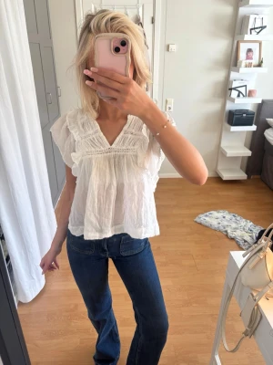 Vit blus med volanger Neo Noir   - Söt vit blus med v-ringning och volangdetaljer längs axlar och bröst. Blusen har korta ärmar och är gjord i ett tunt, lätt material med broderade detaljer. 
