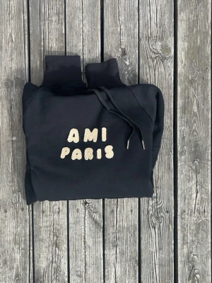 Svart hoodie med AMI PARIS-tryck - Säljer en svart hoodie från AMI PARIS med fluffigt vitt texttryck på bröstet. Hoodien har huva med dragsko och ribbade muddar vid ärmslut och nederkant. Perfekt för dig som gillar streetwear och vill ha något stilrent men ändå unikt.