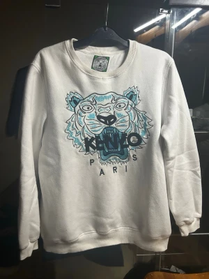 Vit Kenzo sweatshirt - Säljer en vit sweatshirt från Kenzo med det ikoniska tigerhuvudet broderat i blått och svart på bröstet. Tröjan har rund halsringning och ribbade muddar vid ärmslut och nederkant. Perfekt statement-plagg för dig som gillar streetwear.