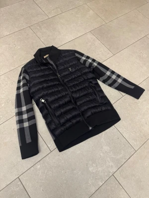 Svart Burberry cardiganjacka med rutigt mönster - Snygg svart Burberry cardiganjacka med quiltad front och ärmar i stickat material med grått rutmönster. Jackan har dragkedja framtill, två fickor med dragkedja och ribbade muddar. Perfekt för dig som gillar en modern och stilren look.