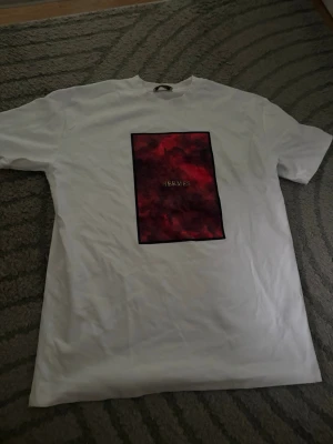 Vit t-shirt med röd mönstrad ruta - Säljer en vit t-shirt med en stor rektangulär ruta på bröstet i röd och mörk ton' i mitten. T-shirten har korta ärmar och en klassisk passform. Perfekt för dig som vill sticka ut med en unik detalj.