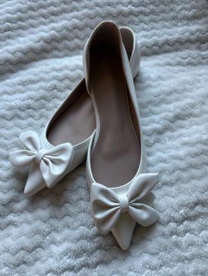 Vita ballerinaskor med rosett - Stilrena vita ballerinaskor med spetsig tå och stora dekorativa rosetter framtill. Skorna har en slät ovansida i ett glansigt material som liknar satin och en ljus, räfflad sula för bättre grepp. Perfekta för dig som vill ha en söt och elegant look. Helt nya och aldrig använda som ni ser på bilden 
