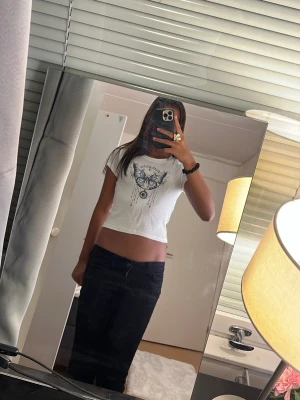 Vit t-shirt med fjärilstryck från Lindex - Snygg vit t-shirt från Lindex med ett stort tryck av en blå och svart fjäril och texten 'dream catcher' på bröstet. T-shirten har korta ärmar och rund halsringning. Perfekt för dig som gillar coola tryck och vill sticka ut lite extra.