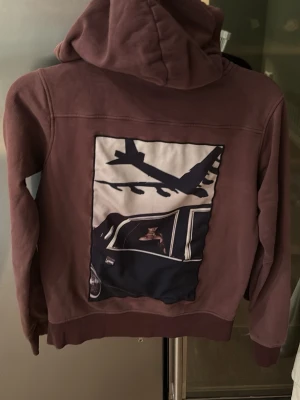 Limitato hoodie - Säljer en brun limitato hoodie med huva och dragkedja framtill. På ryggen finns ett stort tryck med motiv av ett flygplan och en bil. Hoodien har två fickor med dragkedja och ribbade muddar. Perfekt för dig som gillar unika detaljer.