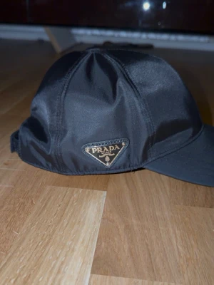 Svart keps från Prada i nylon - Snygg svart keps från Prada i glansigt nylonmaterial. Klassisk form med böjd skärm och justerbar baksida. På sidan sitter Pradas ikoniska metallogo i trekant. Perfekt accessoar för en trendig och stilren look.