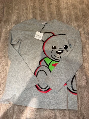 Grå långärmad topp med björntryck moschino - Snygg grå långärmad topp med ett stort tryck av en tecknad björn i svart, rött och grönt framtill. Toppen har rund hals och är tillverkad i mjukt material som känns skönt mot huden. Perfekt för dig som gillar lekfulla och färgglada motiv.