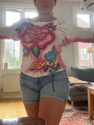 Vit topp med stora blommor Twinset - Säljer en vit topp från Twinset med stora broderade blommor i rosa, blått och grönt framtill. Tröjan har rosa detaljer längs sidorna och ärmarna samt paljetter på blommorna för extra effekt. Perfekt för dig som gillar färg och unika plagg.made in italy st S men passar också M