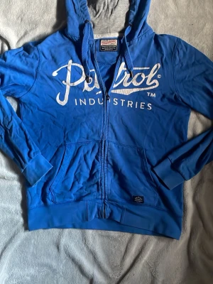 Blå zip hoodie från Petrol Industries - Snygg blå hoodie med dragkedja från Petrol Industries. Stor vit logga framtill och två stora fickor. Mjuk bomullskänsla och huva med snörning. Perfekt för en avslappnad streetwear-look.