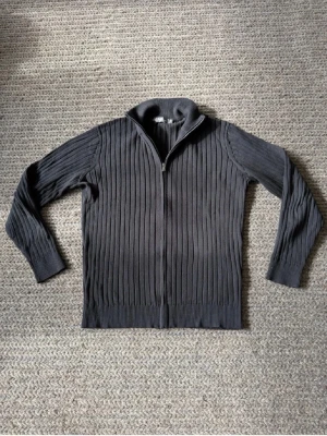 Svart ribbstickad ziptröja - Säljer en svart ribbstickad tröja från Dressmann med dragkedja framtill och hög krage. Tröjan har långa ärmar och är tillverkad i ett mjukt, stretchigt material som passar perfekt för kyligare dagar. Enkel och stilren design som funkar till det mesta.
