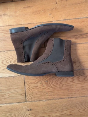 Bruna Chelsea boots i mocka från Arezzo - Snygga Chelsea boots i mörkbrun mocka med elastiska sidopaneler och dekorativa broguemönster längs sömmarna. Bra skick, storlek 42, inga defekter ( små veck i fram ). Hör av er vid funderingar.