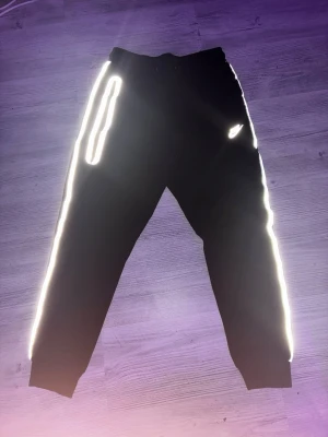 Svarta Nike mjukisbyxor med reflex - Säljer ett par svarta mjukisbyxor från Nike med feta reflexdetaljer längs sidorna och logga på låret. Byxorna har resår i midjan och vid bensluten samt fickor på sidorna. Perfekta för chill eller streetwear.