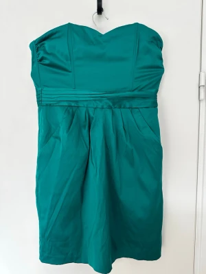 Grön axelbandslös kort klänning - Snygg grön axelbandslös klänning med hjärtformad urringning och veckad midjedetalj. Klänningen har en elegant, enkel design och är tillverkad i ett glansigt material som ger en lyxig känsla. Perfekt för dig som vill sticka ut med färg.