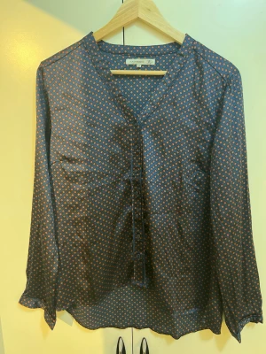 Mönstrad blus från Flash Woman - Snygg blus från Flash Woman i storlek M. Blusen är mörkblå med små orangea diamantmönster, har V-ringning och knappar framtill. Tillverkad i ett tunt, glansigt material som känns skönt mot huden. Perfekt för dig som gillar stilrena mönster.