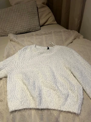 Vit fluffig stickad tröja Divided - Mysig vit stickad tröja från Divided by H&M. Tröjan har en fluffig struktur, rund halsringning och långa ärmar. Perfekt för kalla dagar när du vill vara både bekväm och snygg. Materialet känns mjukt och gosigt mot huden.