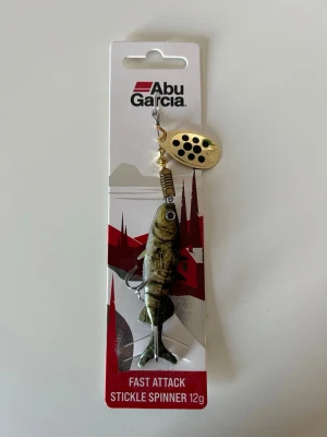 Abu Garcia Stickle Spinner 12g - Fiskedrag från Abu Garcia, modell Fast Attack Stickle Spinner på 12g. Draget har en naturtrogen fiskform i grönt och brunt med svarta detaljer samt ett guldigt spinnblad med svarta prickar. Perfekt för sportfiske och predatorfiske.
