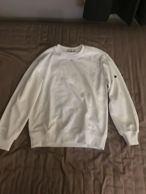 Vit sweatshirt från Stone Island - Säljer en clean vit sweatshirt från Stone Island med klassisk rund hals och ribbade muddar. Tröjan har den ikoniska svarta patchen med gult och grönt broderi på vänster ärm. Perfekt för dig som gillar streetwear och enkel stil.