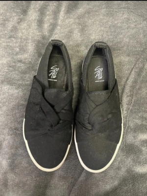 Svarta slip-on sneakers med knut - Svarta slip-on sneakers från So All med snygg knutdetalj på ovandelen. Skorna har en vit platåsula och är tillverkade i tygmaterial. Perfekta för en chill och trendig look.
