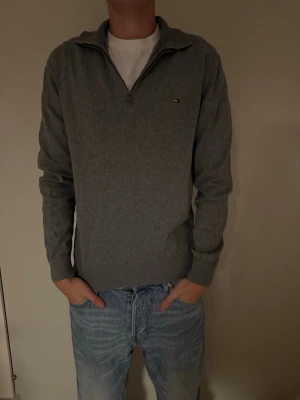 Grå halv-zip tröja från Tommy Hilfiger - Snygg grå långärmad half-zip tröja från Tommy Hilfiger med halv dragkedja och liten logga på bröstet i storlek L. Tröjan har ribbade muddar och en fungerande dragkedja med krage som kan bäras både uppvikt och nedvikt. Perfekt till jeans för en clean och stilren look.