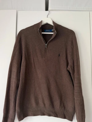 Brun halvzip tröja från Polo Ralph Lauren - Snygg brun stickad tröja från Polo Ralph Lauren med halv dragkedja och diskret logga på bröstet. Tröjan har ribbade muddar vid ärmslut och nederkant samt hög krage. Perfekt för lager på lager och en klassisk stil.