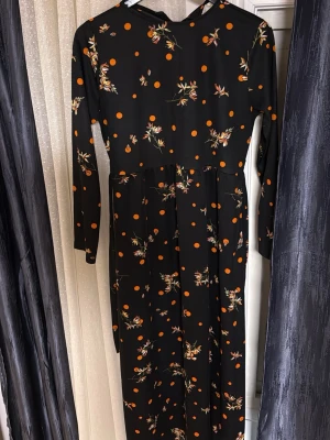 Svart maxiklänning med orange prickar - Lång svart maxiklänning med orange prickar och blommönster. Klänningen har långa ärmar och rund halsringning. Tyget ser ut att vara mjukt och följsamt, perfekt för dig som gillar en unik och färgglad stil.