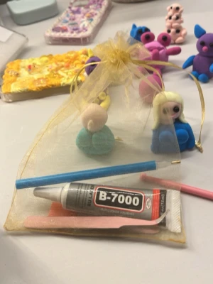 Verktygskit till blondi  - Ett DIY pysselkit i en guldfärgad organzapåse. Innehåller färgglad lera, B-7000 lim, två verktyg i blått och rosa samt små figurer i olika färger. Perfekt för att skapa egna dekorationer eller figurer.
