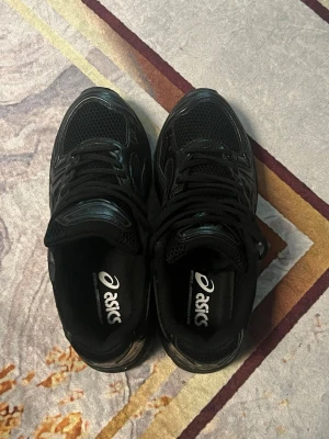 Svarta Asics sneakers - Säljer ett par helsvarta Asics sneakers med klassisk mesh och syntetpaneler. Skorna har en sportig look med tydliga Asics-loggor på sidan och tungan. Lågt skaft och snörning för skön passform. Perfekta för dig som gillar stilrena och bekväma sneakers.