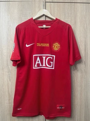 Manchester United 2007/08 Ronaldo 7 - Röd Manchester United matchtröja från Nike med tryck 'Ronaldo 7' på ryggen och AIG-logga på bröstet. Tröjan har korta ärmar, klubbmärke och detaljer från Champions League-finalen 2008. Perfekt för fotbollsfans!